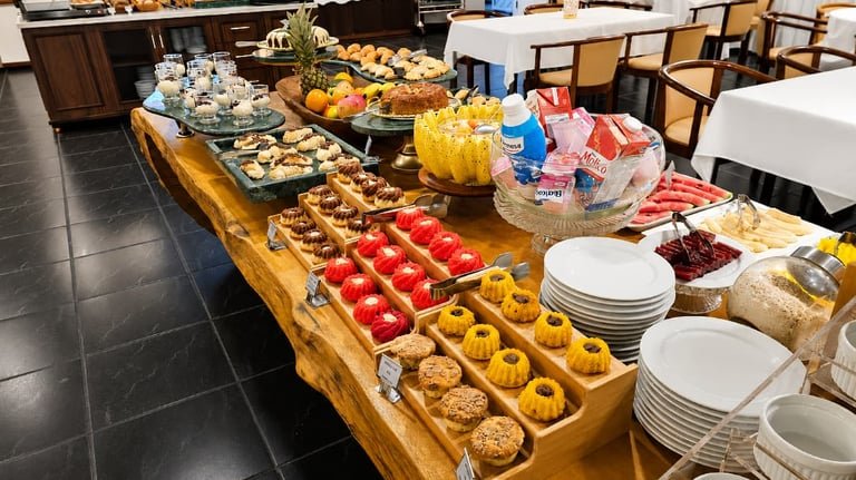 Mesa de café do Hotel Santoni com pequenos bolos, frutas e louças, junto às mesas do salão